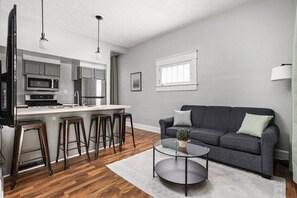 A modern kitchen with a microwave, stove, and bar stools. There is a gray sofa with a small table and a potted plant. - The Sea Turtle - Private Studio, Walk to Mass Ave (Indianapolis)