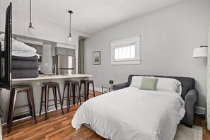 A bedroom with a bed, a small desk, stools, a window, and a kitchenette. - The Sea Turtle - Private Studio, Walk to Mass Ave (Indianapolis)