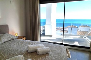 A hotel room with a bed, a nightstand with a lamp, and a large window offering a view of the sea and buildings. - Sea View Apartment in La Paloma de Manilva (Manilva)