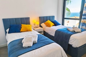 Ein Hotelzimmer mit zwei Betten, jeweils mit blauer Bettwäsche und gelben Kissen, einem Nachttisch mit einer Lampe sowie Blick auf das Meer durch ein großes Fenster.