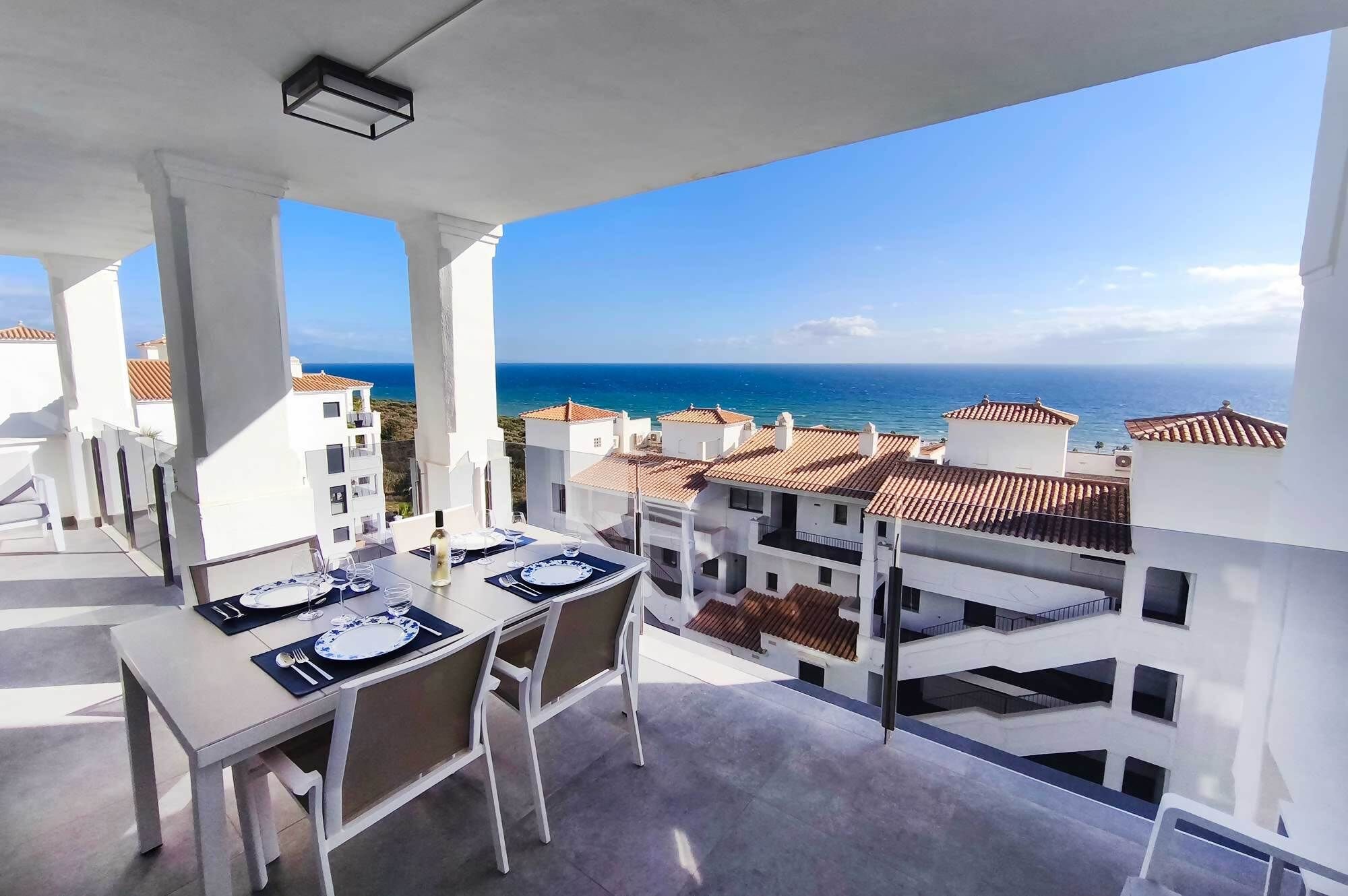 A balcony with a table set for dining, overlooking a coastal area with buildings and the sea.