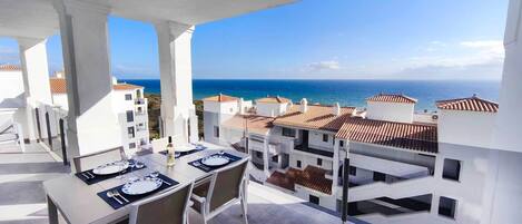 A balcony with a table set for dining, overlooking a coastal area with buildings and the sea.