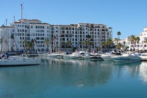 A marina with multiple boats docked, a large building with balconies, and palm trees in the background. - Apartment 'Apartamento con terraza y vistas al mar (Castillo De La Duquesa)