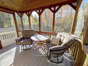 A covered outdoor seating area with a table and two chairs, surrounded by wooden railings and a view of trees. - Kennebec River Cabin with Barrel Sauna & Porch (Richmond)