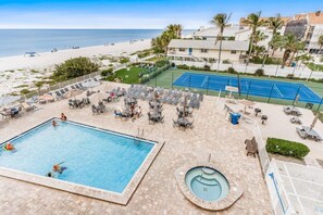 A pool area with a tennis court, lounge chairs, and a view of the beach. - Reef Club 508 - Beachfront 3br/2ba luxury condo with pool and tennis (Indian Rocks Beach)