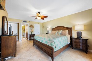 A bedroom with a bed, a nightstand, a dresser, a ceiling fan, and a bathroom visible through an open door. - Reef Club 508 - Beachfront 3br/2ba luxury condo with pool and tennis (Indian Rocks Beach)
