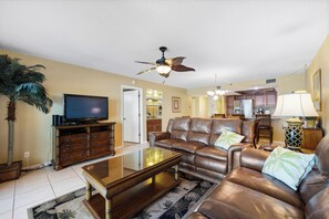 A living room with a leather sofa, a coffee table, a television, and a ceiling fan. - Reef Club 509 - Beachfront 3br/2ba luxury condo with pool and tennis (Indian Rocks Beach)