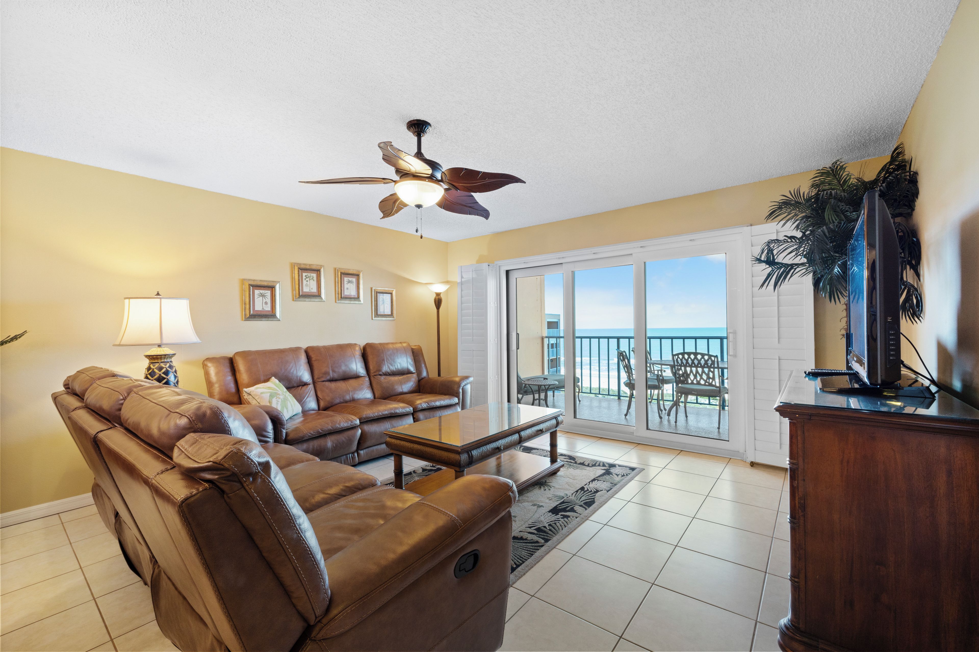 A living room with a brown leather sofa, a coffee table, a ceiling fan, and a view of the ocean through a sliding glass door.