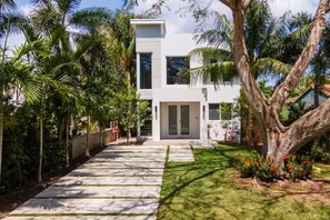 Une maison moderne à deux étages, dotée d’une allée pavée et entourée d’une végétation luxuriante et de palmiers.