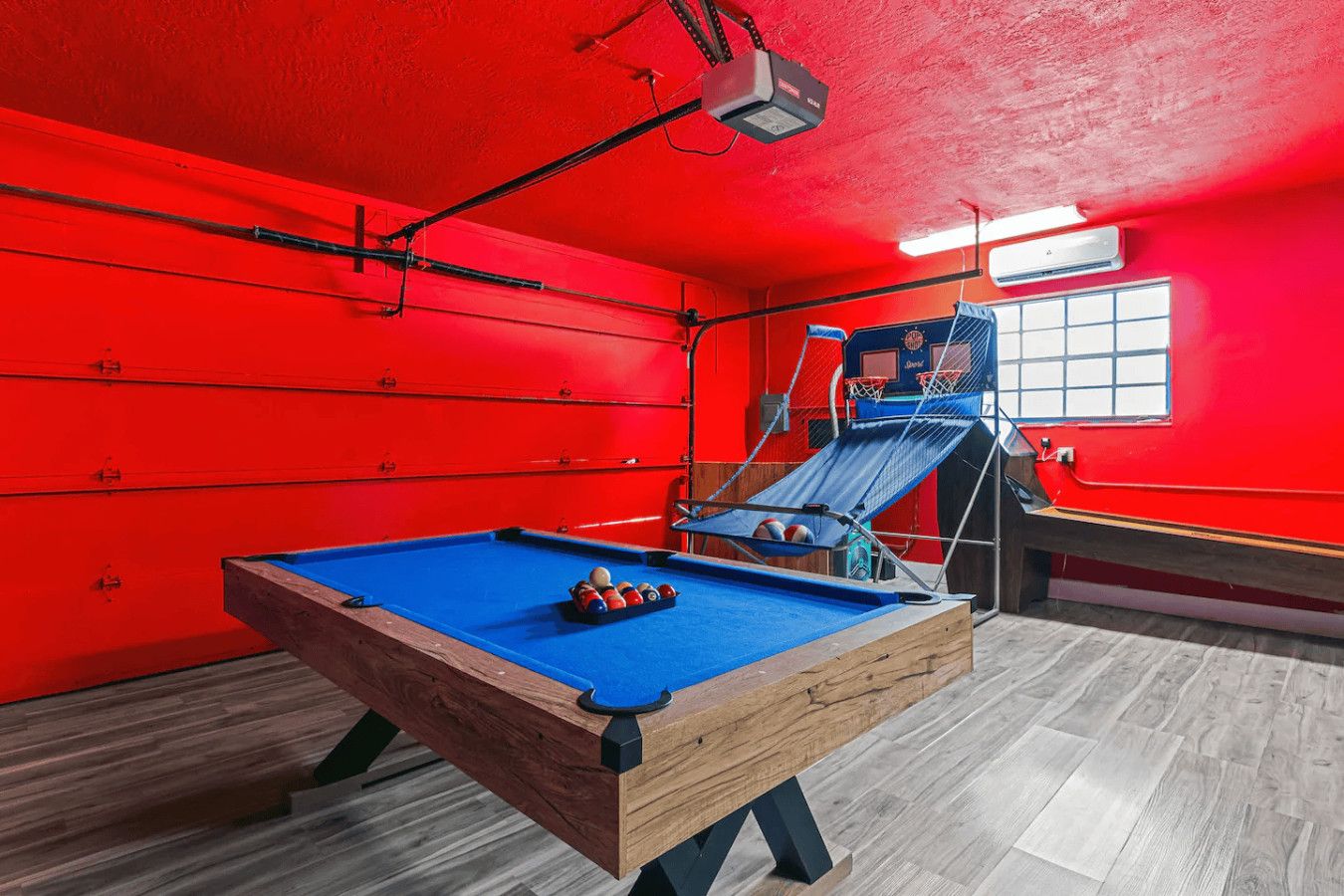 A pool table with balls, a projector, and a basketball hoop in a room with red walls and wooden flooring.