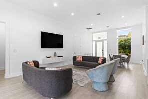 Sala de estar moderna com televisão de tela plana, sofás cinza, área de jantar e vista para o verde lá fora.