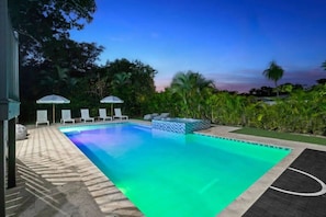 A rectangular swimming pool with a tiled edge, surrounded by lounge chairs and umbrellas, set against a backdrop of trees and a clear sky. - Modern Luxe 2021Built Heated Pool/Jacuzzi/Gameroom (Fort Lauderdale)