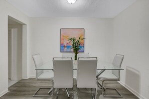 Un comedor moderno con una mesa de vidrio y sillas blancas, un cuadro en la pared y una planta en maceta.