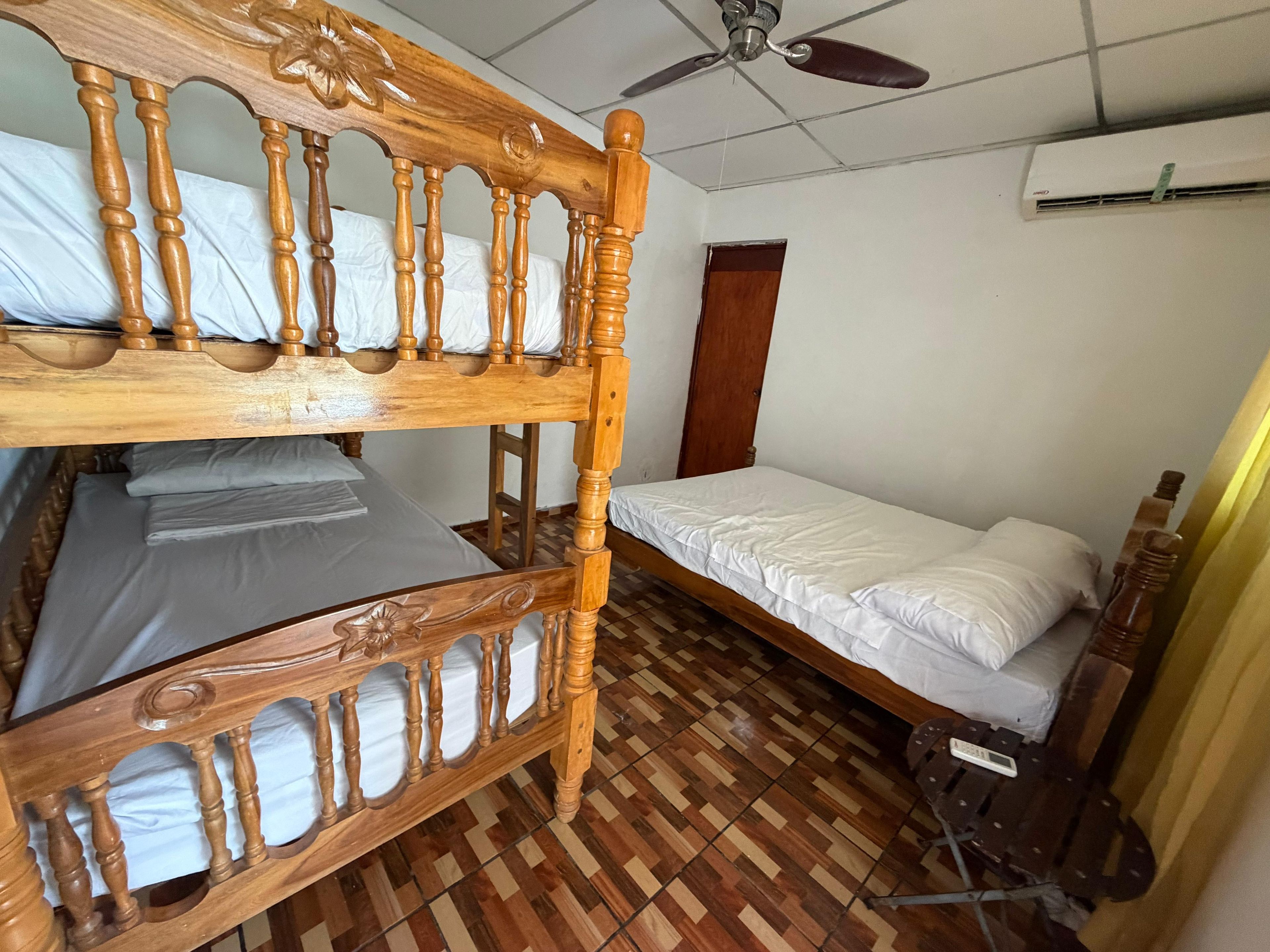 A room with a bunk bed, a single bed, a ceiling fan, an air conditioning unit, and a wooden floor.