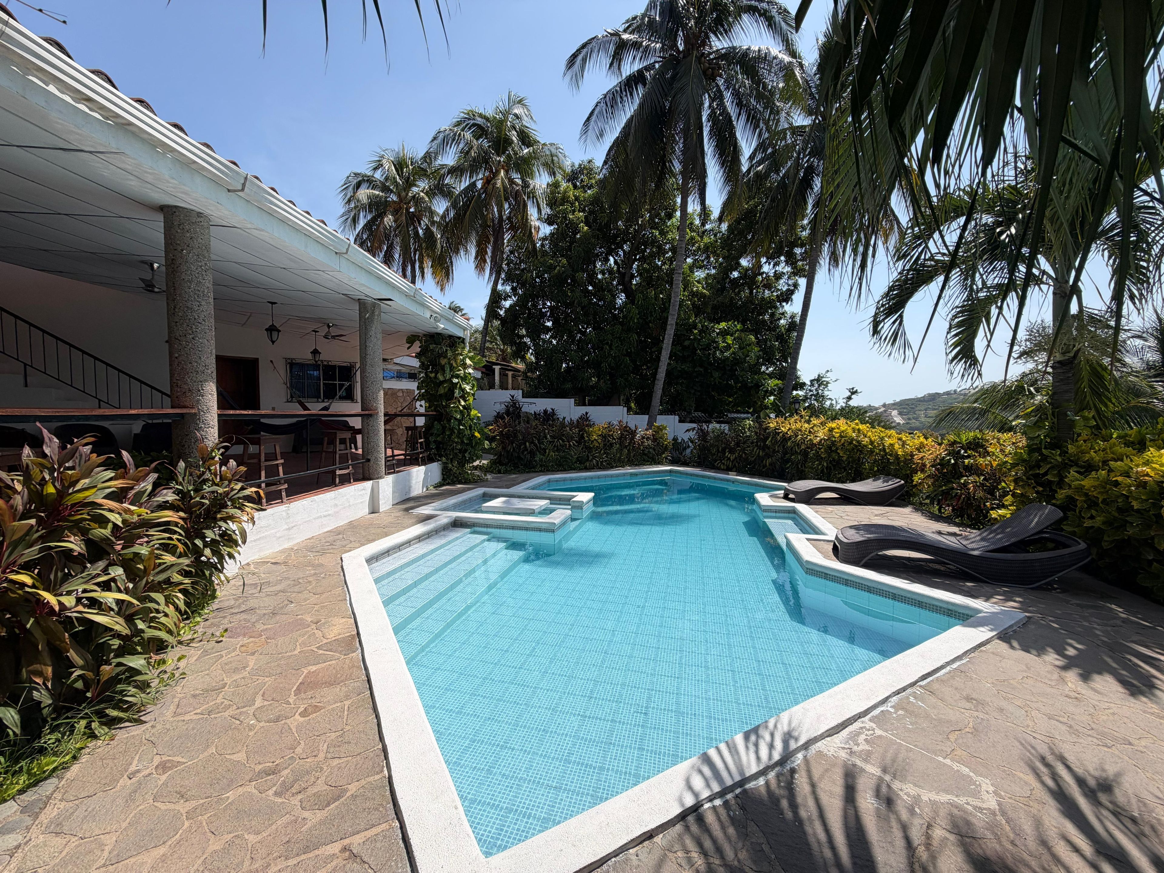 A swimming pool with a tiled edge, surrounded by palm trees and a covered seating area.