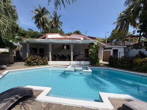 Ein Swimmingpool mit überdachter Terrasse, Palmen und einem Haus mit rotem Dach.