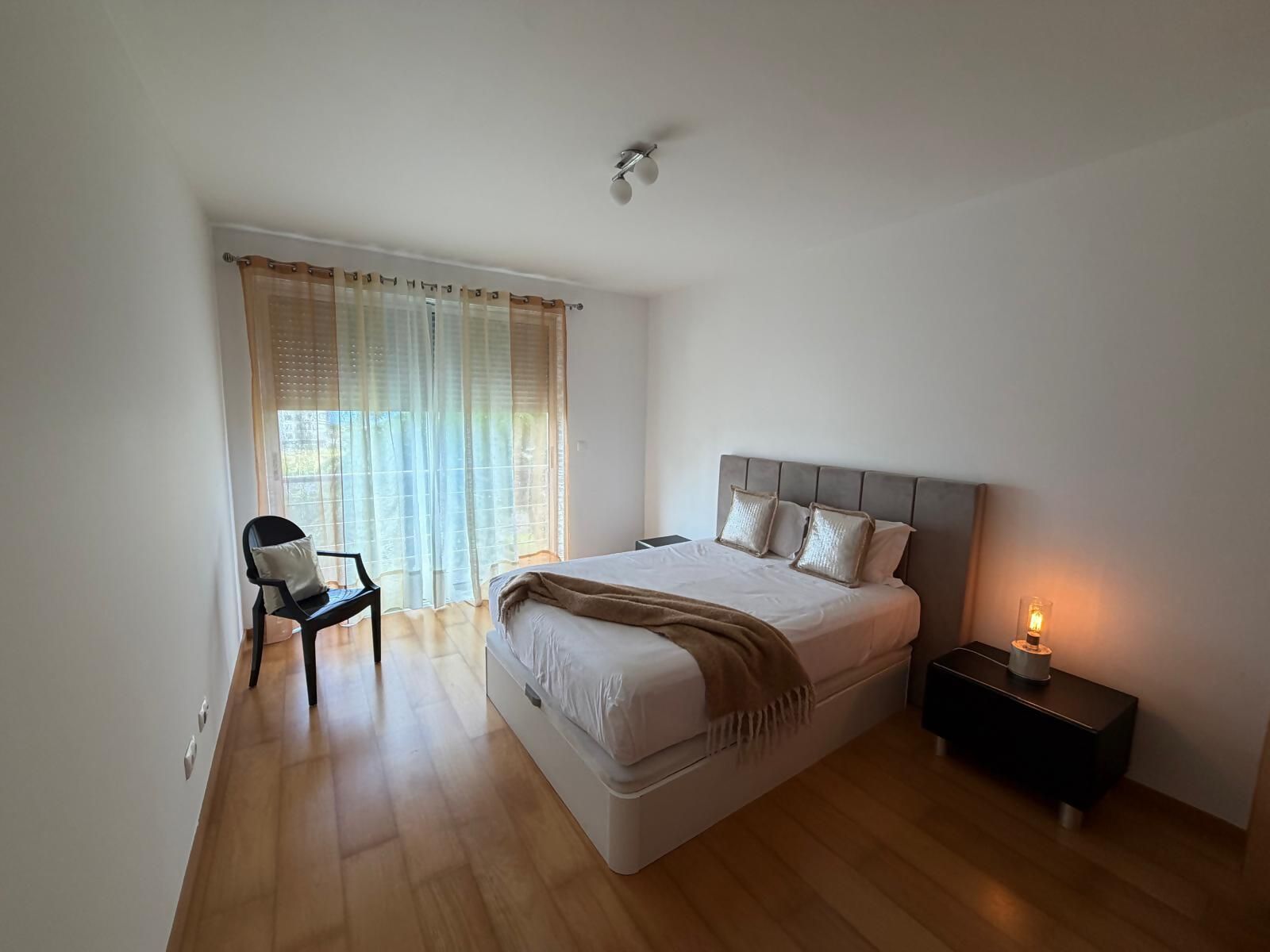 A bedroom with a bed, bedside table, chair, and a window with curtains.