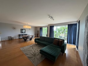 A modern living room with a green sofa, a dining table with chairs, and a large window with curtains. - AZORES APARTMENTS PAIM 1NASC (Ponta Delgada)