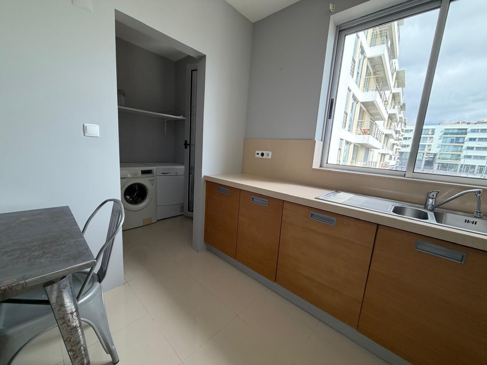 A compact kitchen with a built-in washing machine, wooden cabinets, and a window with a view of buildings.