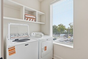 A laundry room with a front-loading washer and dryer, a shelf with folded towels, and a window with blinds. - Pretty in Pink: Rooftop Hot Tub & Sauna | 4 Ensuite Bedrooms | Sleeps 14 (Nashville)