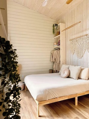 A bedroom with a wooden bed frame, a macrame wall hanging, and a plant. - Grace Nature Retreat•Shine & Rise Wellness (Sainte-Marie-de-Kent)