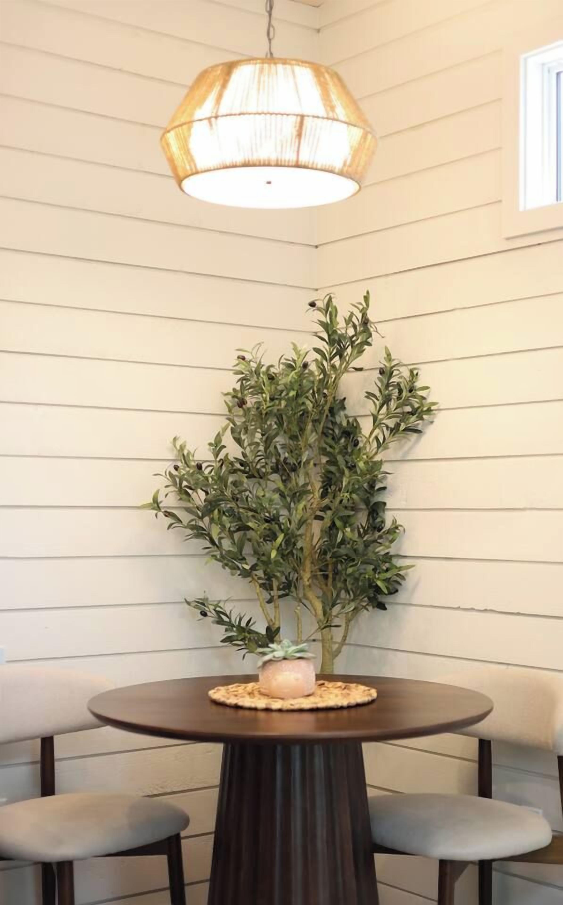 A round dining table with a dark wood finish, two grey cushioned chairs, a potted plant with green foliage, and a hanging light fixture with a woven shade.