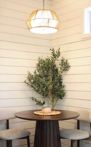 A round dining table with a dark wood finish, two grey cushioned chairs, a potted plant with green foliage, and a hanging light fixture with a woven shade. - Grace Nature Retreat•Shine & Rise Wellness (Sainte-Marie-de-Kent)