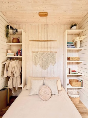 A room with a bed, a macrame wall hanging, shelves with books and baskets, and a wooden ceiling.