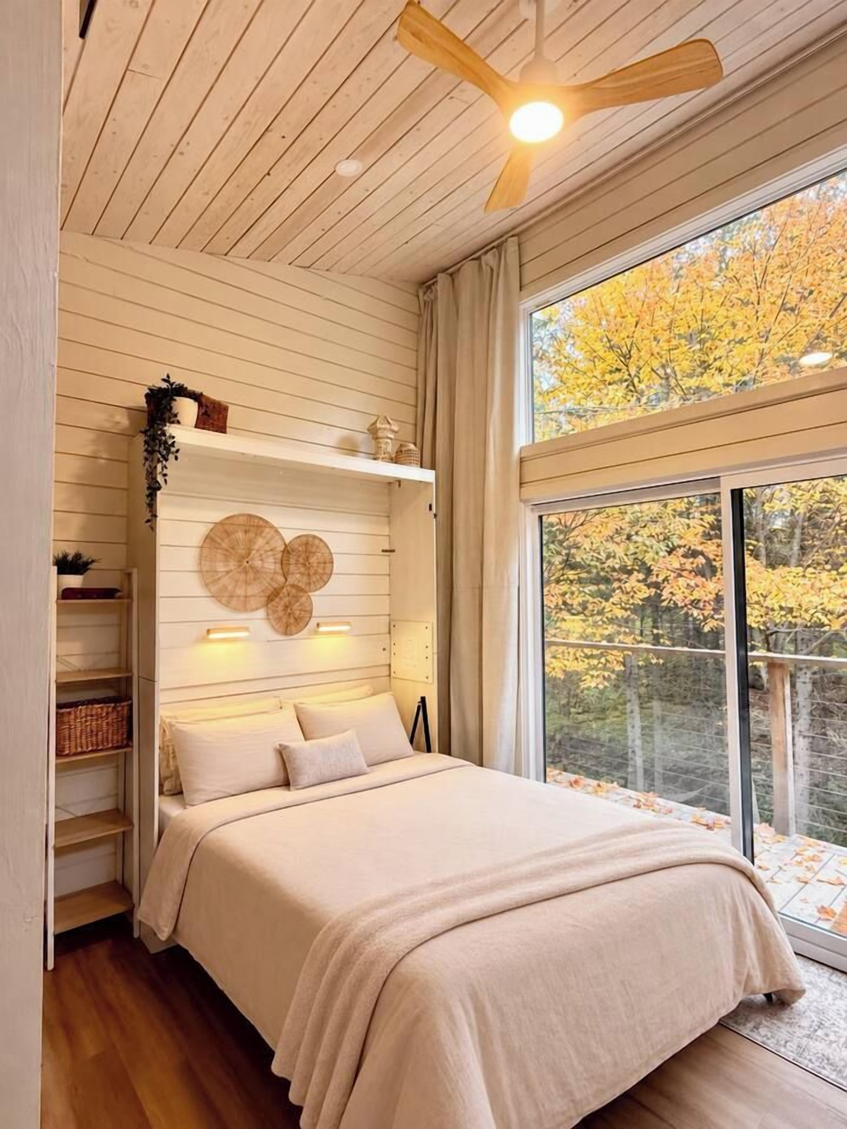 Una camera da letto con un letto, una ventola a soffitto, una finestra vista alberi e mensole con oggetti.
