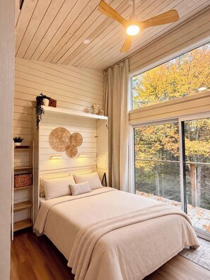 A bedroom with a bed, a ceiling fan, a window with a view of trees, and shelves with items. - Haven Nature Retreat•Shine & Rise Wellness (Sainte-Marie-de-Kent)