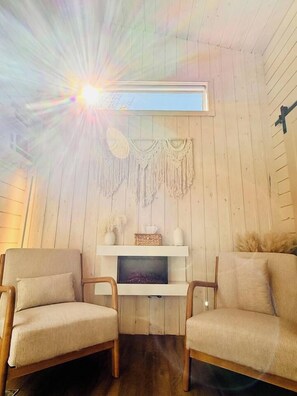 A room with wooden walls, two beige armchairs, a white shelf with decorative items, and a macrame wall hanging. - Haven Nature Retreat•Shine & Rise Wellness (Sainte-Marie-de-Kent)