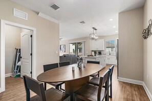 Een moderne keuken met witte kastjes, een eethoek met een houten tafel en stoelen, en een zichtbaar raam met jaloezieën.
