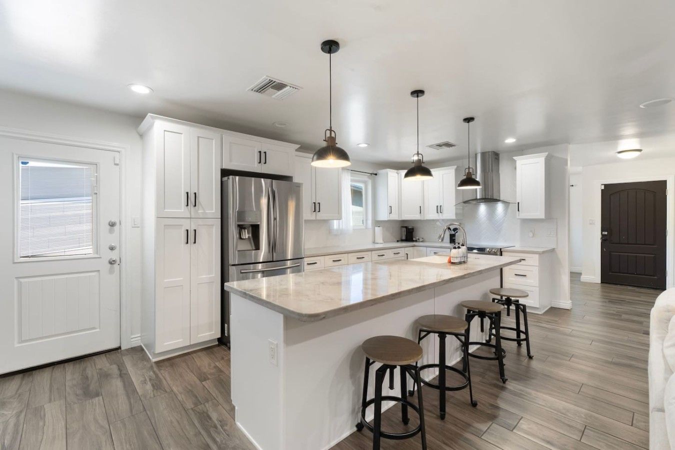 A modern kitchen with white cabinetry, a stainless steel refrigerator, a large island with stools, pendant lighting, and a dark wooden door.