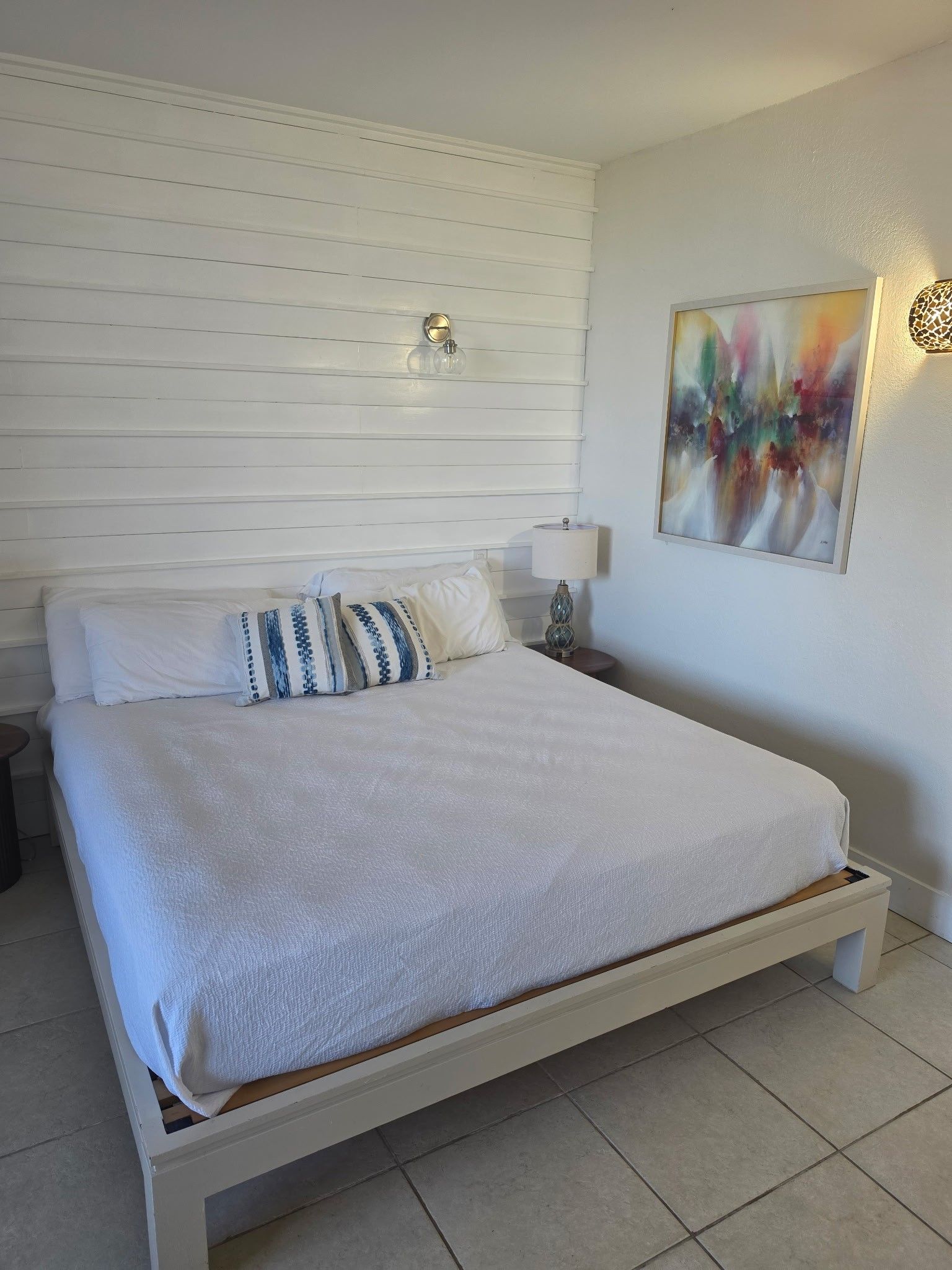 Un dormitorio con cama, almohadas, lámpara de noche, lámpara de pared y un cuadro.