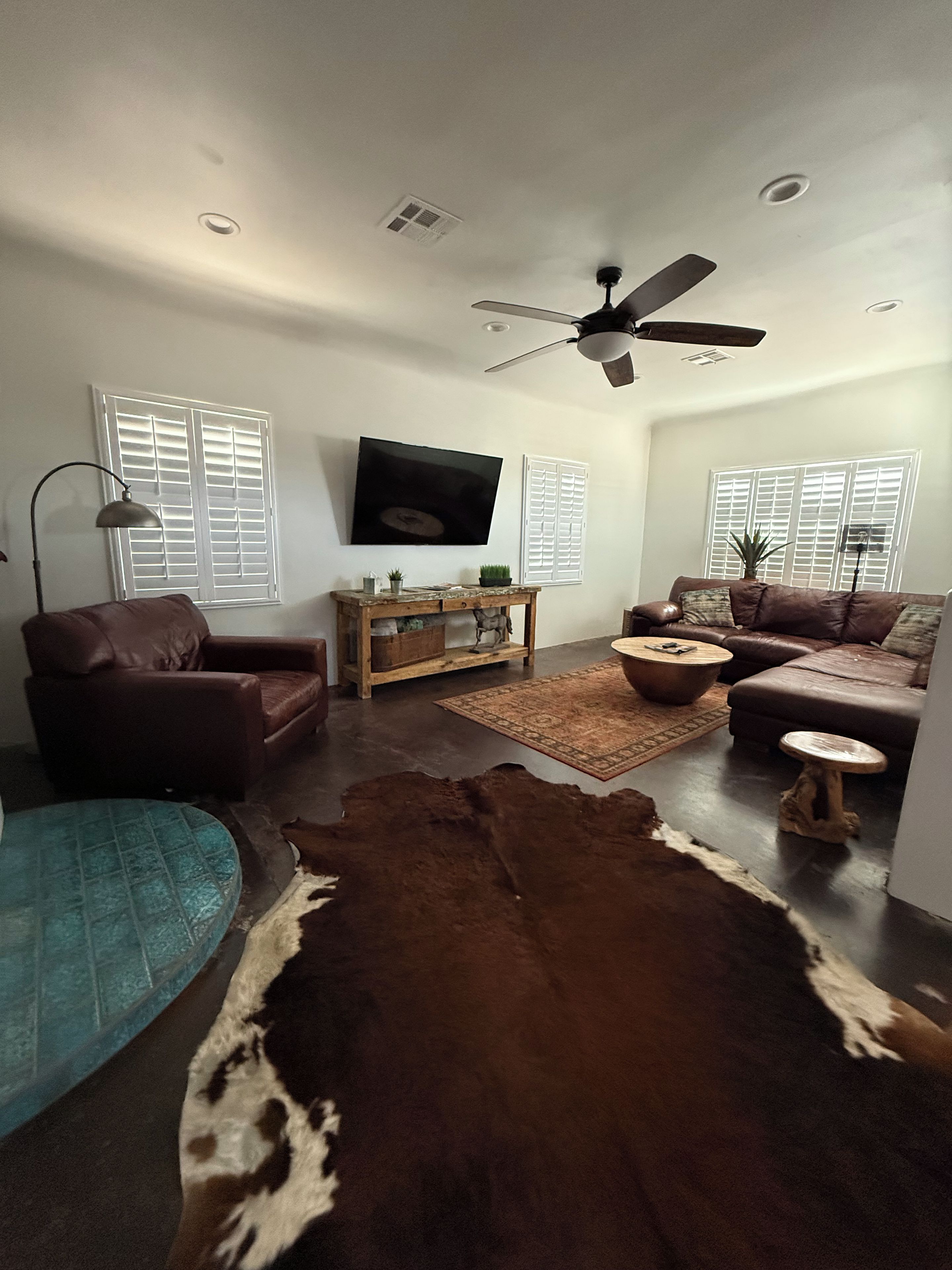 A living room with a brown leather sofa, a wooden coffee table, a television mounted on the wall, and a ceiling fan.
