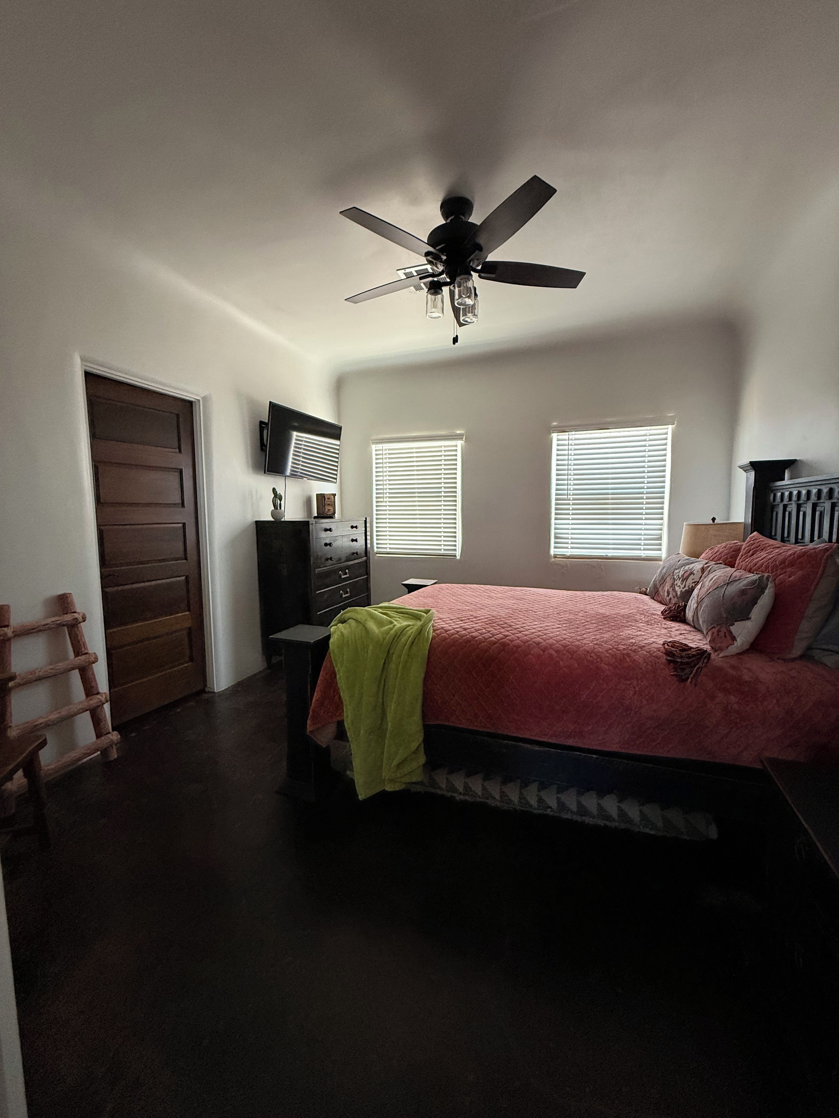 A bedroom with a bed, a ceiling fan, a dresser, a TV, and two windows with blinds.