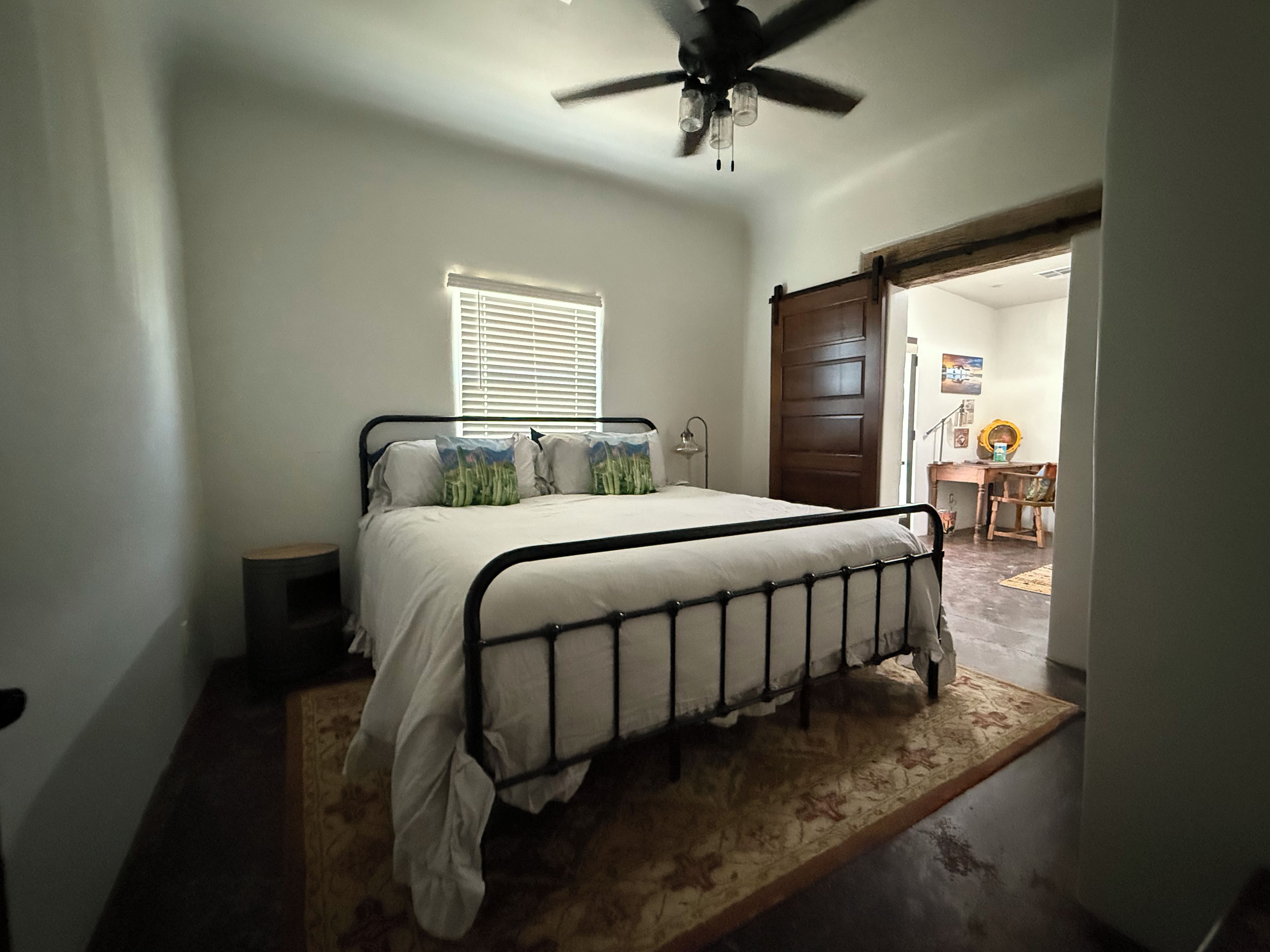 A bedroom with a bed, a ceiling fan, a window with blinds, and a door leading to another room.