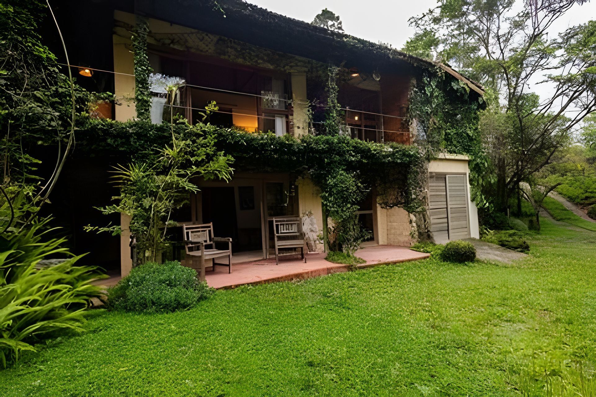 Um edifício de dois andares com varanda, rodeado por vegetação exuberante e um gramado bem cuidado.