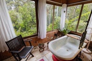 A bathroom with a large bathtub, wooden bench, and a view of a forest through the window. - Macondo uma Vila (Tapirai)