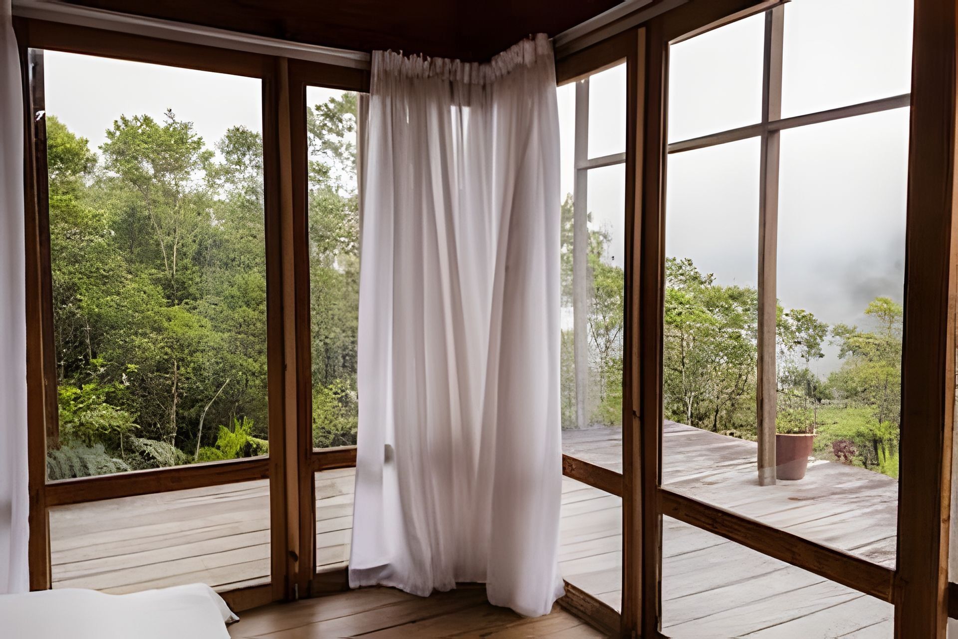 Quarto com janelas amplas, piso de madeira e vista para uma área arborizada.