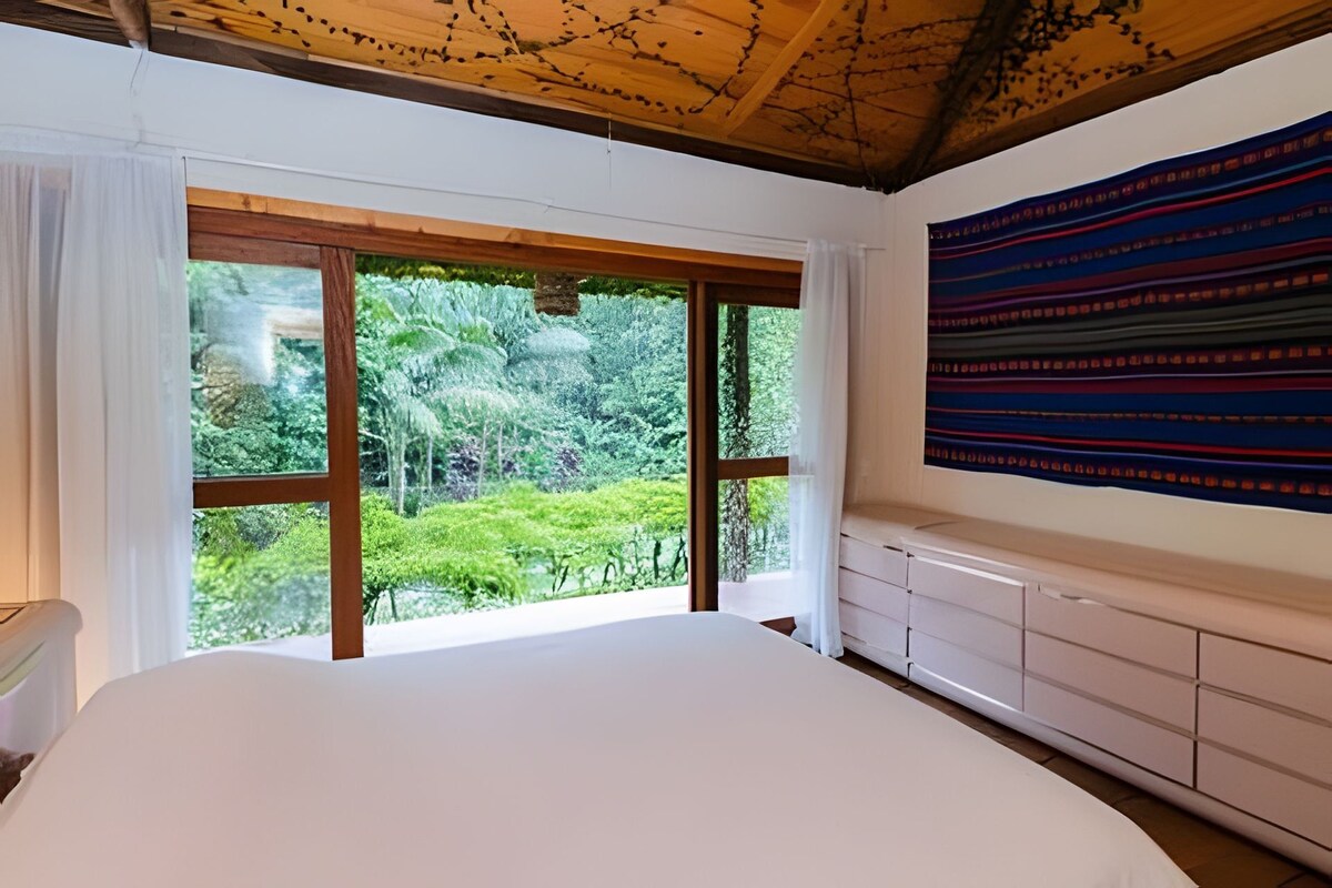 Un dormitorio con una cama grande, vistas a la vegetación a través de ventanas de gran altura y un tapiz colorido en la pared.