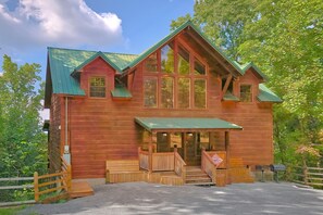 A two-story log cabin with a green roof, large windows, and a front porch. - Mountain Bearadise | Theater, Hot Tub, Game Room, Sleeps 12, Fireplace + Free Attraction Tickets (Sevierville)
