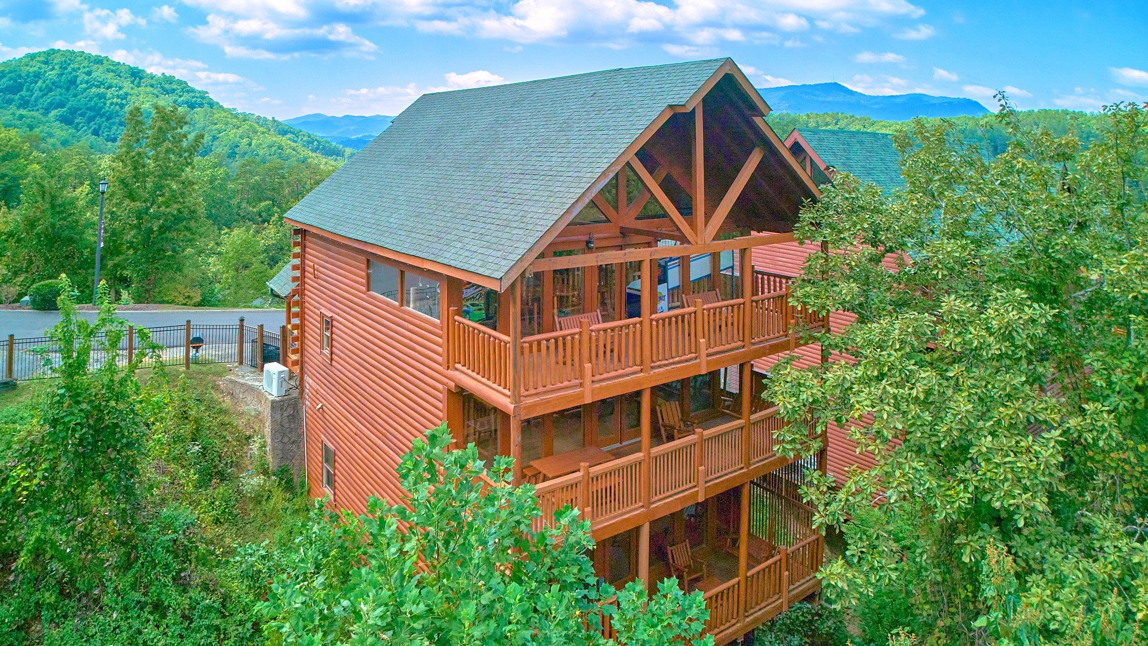 A two-story log cabin with a balcony, surrounded by lush greenery and mountains in the background.