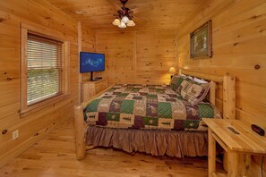 A wooden cabin bedroom with a bed, a television, a window with blinds, and a ceiling fan. - Fun Zone | Theater, Hot Tub, Game Room, Sleeps 12, King Suites, Fireplace + Free Attraction Tickets (Sevierville)