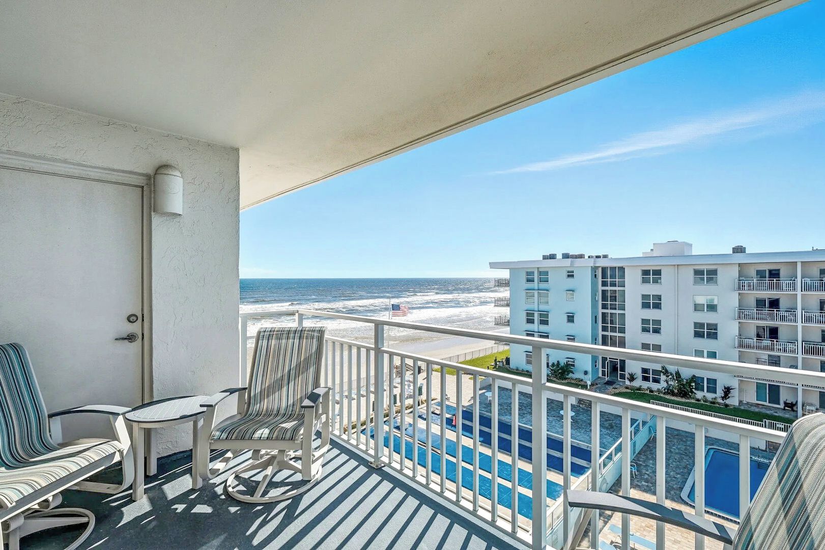 A balcony with a view of the beach and ocean, a pool, and a building with multiple floors.