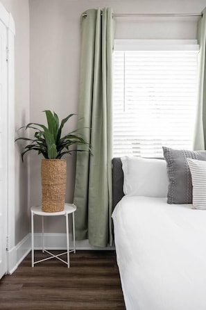 Un dormitorio con cama, una mesita blanca, una planta en maceta y una ventana con cortinas.