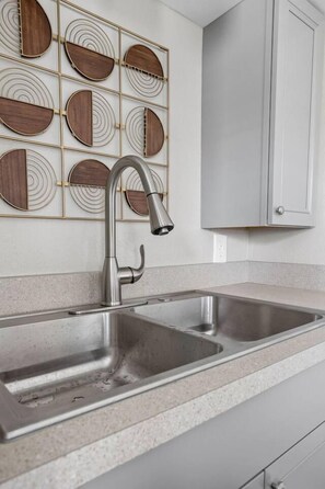 A kitchen with a stainless steel sink, a modern faucet, and a wall-mounted wooden and metal decorative panel. - The Koala - Sunny King Studio -Walk to Mass Ave (Indianapolis)