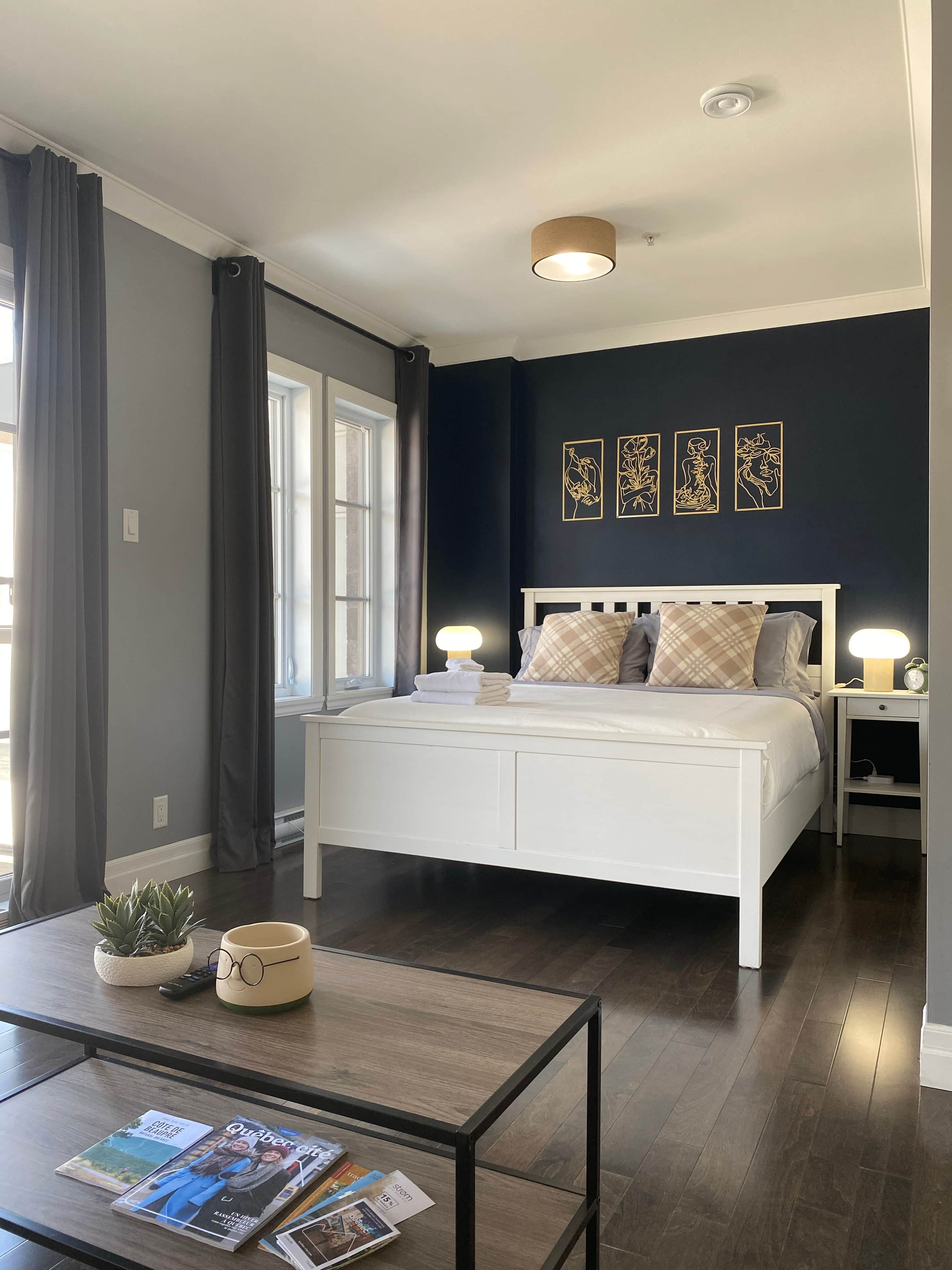 Un dormitorio con cama, lámparas de noche, mesita de noche, ventana con cortinas y una mesa de centro con revistas.