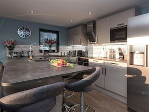 A modern kitchen with a central island, bar stools, stainless steel appliances, and a window with a view. - Whispering Reeds, Hot Tub and Private Quay, Horning (Horning)