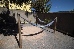 Una hamaca atada entre dos postes de madera sobre un suelo de grava, con un muro de concreto y vegetación al fondo.